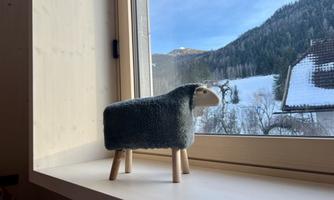 A cute stuffed sheep is standing on a windowsill. Outside, a snowy landscape can be seen.