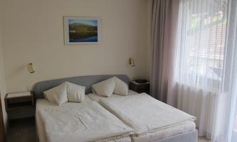 A bright bedroom with two separate beds and white bedspreads. A window with curtains lets in daylight.