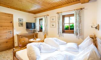 A cozy bedroom with a double bed and many pillows. There is a small table, a television, and windows with a view of greenery.