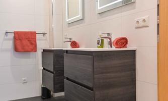 A modern bathroom with two sinks and LED mirrors. The walls are in light tiles, and there is a red towel as well as decorative towels.