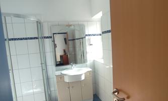 A bright bathroom with a shower, sink, and light tiled floor. The door is open, allowing a view of the room.