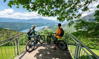 Two cyclists are standing on a lookout platform, enjoying the view of a lake and the surrounding mountains. The sky is clear, and the landscape is green and inviting.