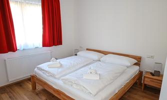 A bright bedroom with a double bed and beautiful red curtains. The floor is wooden and there is a small bedside lamp.