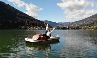 Two people are standing on a boat on a calm lake. In the background, high mountains and green forests are visible.