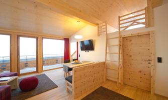 A modern, bright room with wooden paneling and large windows. There is a cozy seating area and a staircase to an upper level.