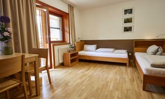 A bright room with two beds and a small table. Large windows let in a lot of light and the furniture is made of wood.