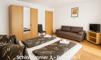 A modern bedroom with two beds, a sofa, and a television. The room features light furniture and a cozy atmosphere.
