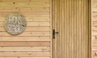 A cozy wooden cabin with a striking door and a sign that reads "HONS STADL". The walls are made of light wooden planks.