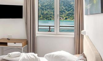 A cozy room with a window that overlooks a beautiful lake and green mountains. In the foreground, there is a bed and a table.