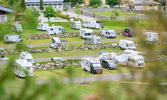 A well-maintained motorhome parking area with several vehicles on green grass. The surroundings are surrounded by trees and buildings.