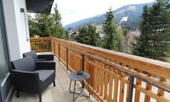 A cozy balcony with two chairs and a table, surrounded by trees and mountains. The view is beautiful and inviting.