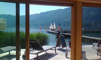 A beautiful view of a calm lake with sailboats in the background. The sun rays illuminate the terrace with loungers.