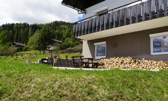 A cozy house with a balcony, surrounded by a green meadow. The woodpile and the trees in the background create a rural atmosphere.