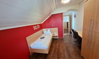 A simply furnished room with a single bed and a red accent wall. There is a table and chairs as well as a wardrobe in the room.