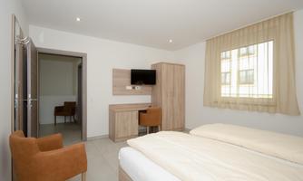 A modern hotel room with two single beds. There is a television, a desk, and a wooden chair as well as a window with bright curtains.