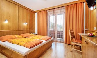 A cozy bedroom with wooden furniture and orange curtains. The room features a double bed and a desk with a fruit bowl.