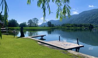 A quiet lake surrounded by green meadows and mountains. A wooden jetty leads into the water.