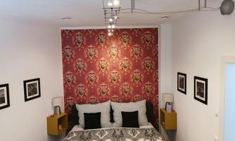 A modern bedroom with a red patterned wall hanging. The bed has an elegant design and there are pictures hanging on the walls.