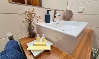 A modern bathroom with a white sink and wooden table. On the shelf are care products and a vase with dried flowers.