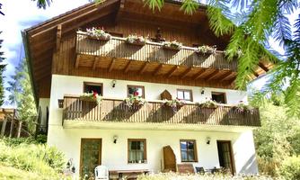 A cozy holiday home with wooden balustrades and flower boxes. It is situated surrounded by a green meadow and trees.