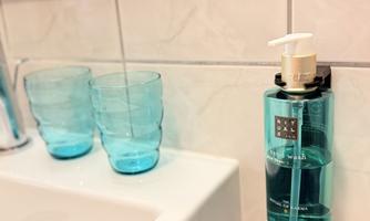A modern bathroom with a white sink and a wall-mounted soap dispenser. Two aqua-blue glasses are placed next to the sink on a wooden shelf.