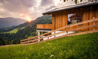 A modern wooden house in a picturesque mountain landscape. The sun sets behind the mountains and the meadows are green and inviting.
