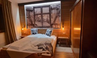 A cozy bedroom with a double bed and warm light. In the background, there is a large window overlooking a winter landscape.