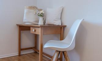 A wooden table with a drawer stands in a corner. Next to it is a white chair and a vase with fresh flowers.