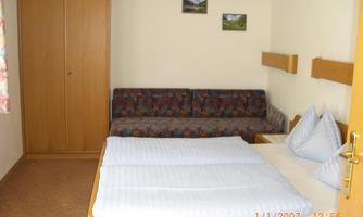 A simply furnished room with a double bed and a couch. Some pictures hang on the wall, and a wardrobe is available.