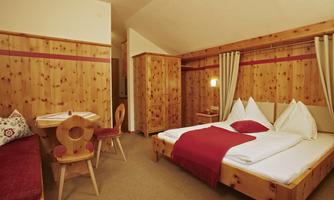 A cozy room with wooden paneling and a double bed. There is a small table and chairs, as well as bright window decorations.