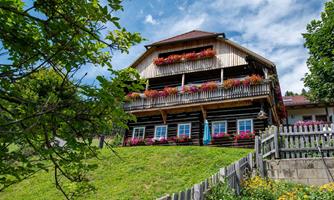 A traditional wooden house with blooming balconies and a well-kept garden. The green meadow and clear sky create an idyllic atmosphere.