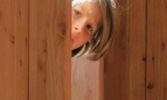 A child is curiously looking through a gap between wooden posts. The light gently falls on the child's face.