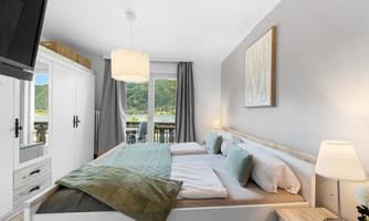 A modern bedroom with a cozy double bed and a view of the mountains. The walls are brightly decorated and there is a large window front with curtains.