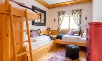 A cozy bedroom with a wooden frame and two beds. Pretty curtains are in front of the window, and there is a colorful wardrobe in the corner.