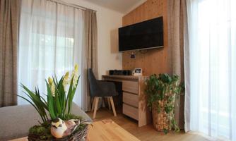 A cozy living area with a television on the wall and a desk. A green plant and a bird decoration give the room an inviting atmosphere.