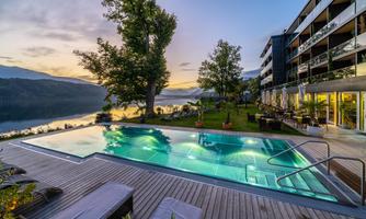 A beautiful outdoor pool overlooking a lake and surrounded by trees. In the background, a modern building stands illuminated in the twilight.