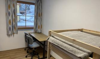 A simple room with a loft bed and a desk. Through the window, you can see snow and a clear sky.