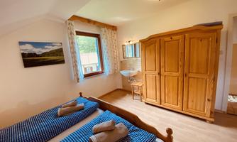 A cozy bedroom with two beds and blue bed sheets. There is a wooden wardrobe and a window with a view outside.