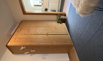 A bright room with a large wooden wardrobe and a view of the kitchen. The floor is covered with a blue carpet and there is a stool with a soft cover.