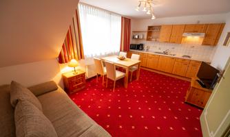 A bright, modern apartment with a cozy seating area and a fully equipped kitchen. The floor is covered with red carpet and large windows provide plenty of light.