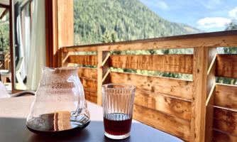 A glass of coffee and a pot are on a table. In the background, green mountains and a blue sky can be seen.