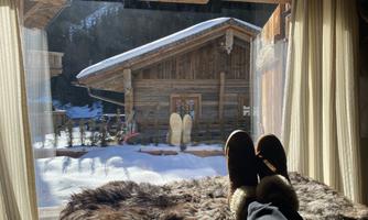 View from a window onto a snowy landscape with a cozy wooden house. A person's feet are resting on a furry surface.