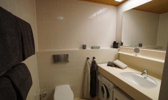 A modern bathroom with white wall tiles and a large mirror. It contains a toilet, washing machine, and towels in various shades of gray.