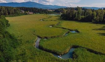 A vast green field with a winding watercourse amidst nature. In the background, trees and mountains are visible.