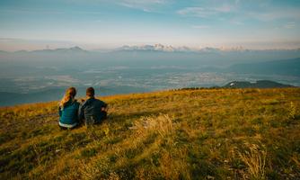 A couple is sitting on a meadow and looking at the mountains in the background. The sky is blue with some clouds, and the landscape is illuminated at sunset.