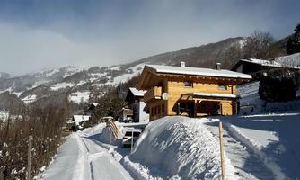 A picturesque winter landscape with snow-covered hills and a wooden house. The path is covered in snow and surrounded by trees.