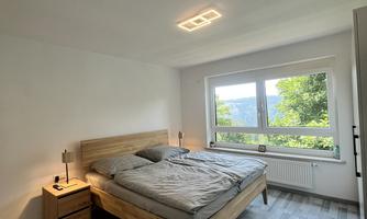 A modern bedroom with a wooden bed and gray bedding. Large windows provide plenty of daylight and a view of the outdoors.