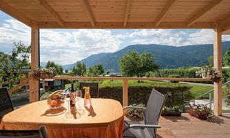 An inviting terrace with a table set with fruit and drinks. In the background, green hills and a cloudy sky can be seen.
