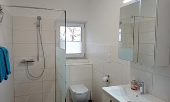 A modern bathroom with a transparent shower and a bright sink. The walls are covered in light tiles, and a window provides natural light.
