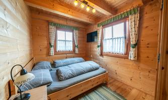 A cozy wooden room with a double bed and two windows. The walls are made of wood, and there is a small table with a lamp.